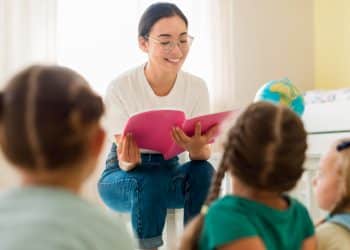 Educadora de óculos sorri sentada ao lado de crianças que desenham em mesa colorida de sala de aula da educação infantil