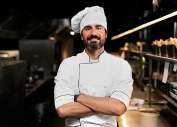 Chef de cozinha com uniforme branco e braços cruzados em cozinha