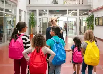 Crianças com mochilas coloridas caminham com professora em corredor escolar, representando a diferença entre escola e colégio.