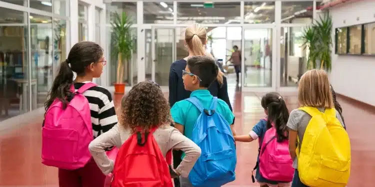 Crianças com mochilas coloridas caminham com professora em corredor escolar, representando a diferença entre escola e colégio.