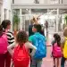 Crianças com mochilas coloridas caminham com professora em corredor escolar, representando a diferença entre escola e colégio.