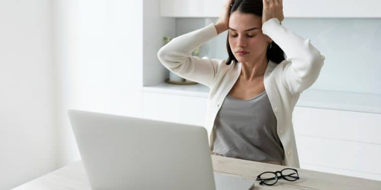 Mulher jovem com casaco bege e blusa cinza segura a cabeça com as mãos diante de notebook e óculos na mesa, simbolizando a dificuldade para manter o foco.