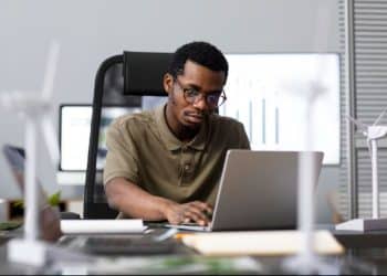 Homem de óculos trabalha concentrado em um notebook em um escritório, com maquetes de turbinas eólicas sobre a mesa