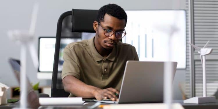 Homem de óculos trabalha concentrado em um notebook em um escritório, com maquetes de turbinas eólicas sobre a mesa