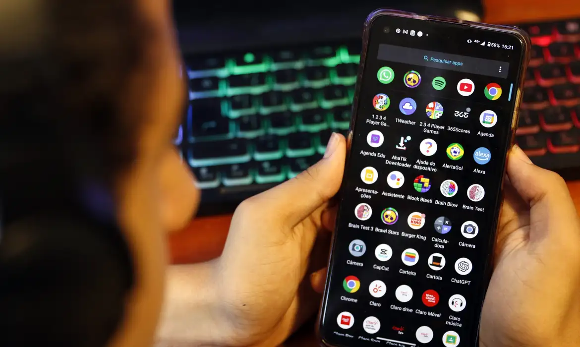 Pessoa segura celular com tela de aplicativos onde aparece o YouTube, ilustrando a nova classificação indicativa para maiores de 16 anos no Brasil