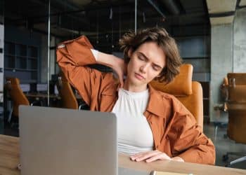 Mulher sentada à mesa de trabalho leva a mão ao pescoço com expressão de cansaço durante uso prolongado do notebook