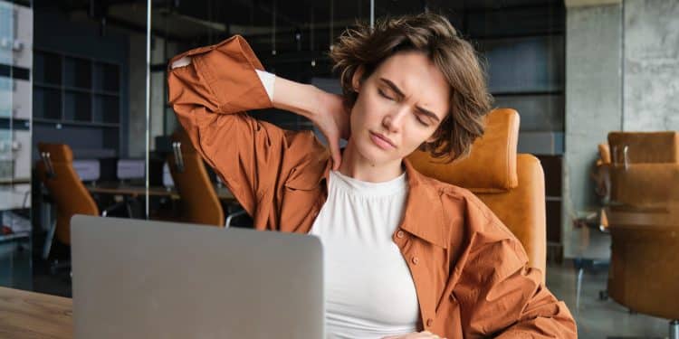 Mulher sentada à mesa de trabalho leva a mão ao pescoço com expressão de cansaço durante uso prolongado do notebook