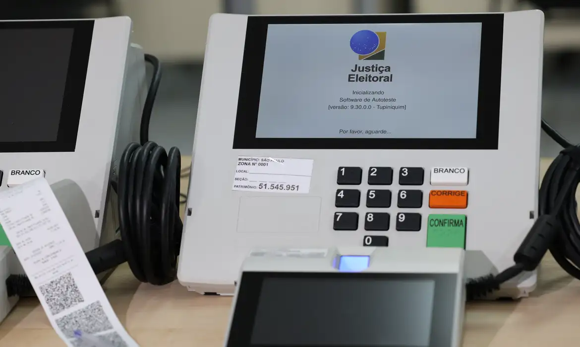 Urnas eletrônicas da Justiça Eleitoral em fase de inicialização para as eleições de 2026, com comprovante de votação visível ao lado do equipamento.