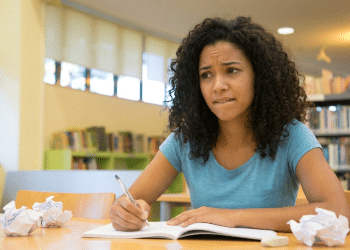 Estudante com expressão de dúvida escreve em um caderno, com papéis amassados sobre a mesa de uma biblioteca.