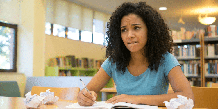 Estudante com expressão de dúvida escreve em um caderno, com papéis amassados sobre a mesa de uma biblioteca.