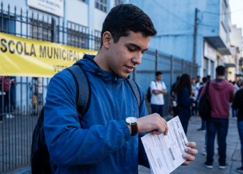 Estudante consulta o relógio e segura o cartão de confirmação em frente a uma escola