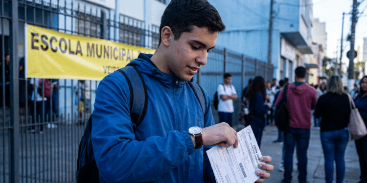 Estudante consulta o relógio e segura o cartão de confirmação em frente a uma escola