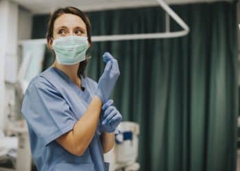 Enfermeira com scrub azul e máscara verde colocando luvas de látex azuis em quarto de hospital com cortina verde, simbolizando a enfermagem hospitalar.