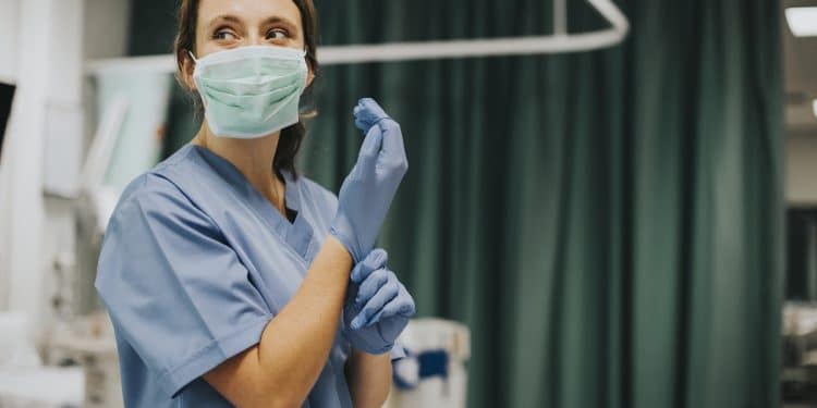 Enfermeira com scrub azul e máscara verde colocando luvas de látex azuis em quarto de hospital com cortina verde, simbolizando a enfermagem hospitalar.