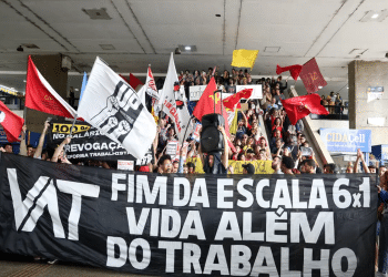 Multidão de manifestantes segura faixa preta com texto "VAT - Fim da Escala 6x1 - Vida Além do Trabalho" e bandeiras vermelhas, simbolizando o fim da escala 6 X 1 e as dúvidas relacionadas a uma possível redução salarial.
