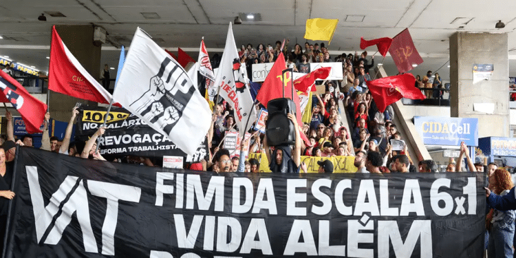 Multidão de manifestantes segura faixa preta com texto "VAT - Fim da Escala 6x1 - Vida Além do Trabalho" e bandeiras vermelhas, simbolizando o fim da escala 6 X 1 e as dúvidas relacionadas a uma possível redução salarial.