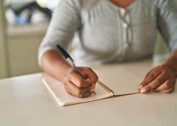 Mãos de mulher escrevendo com caneta preta em caderno espiral sobre mesa branca, blusa cinza, simbolizando o que muda no cérebro quando você anota à mão em vez de digitar no celular.