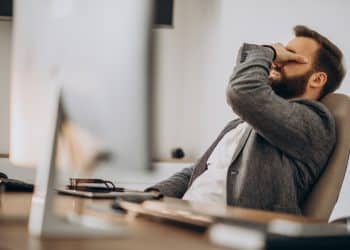 Homem de barba sentado em uma cadeira de escritório com a mão no rosto em expressão de bloqueio mental, representando o esquecimento momentâneo de palavras