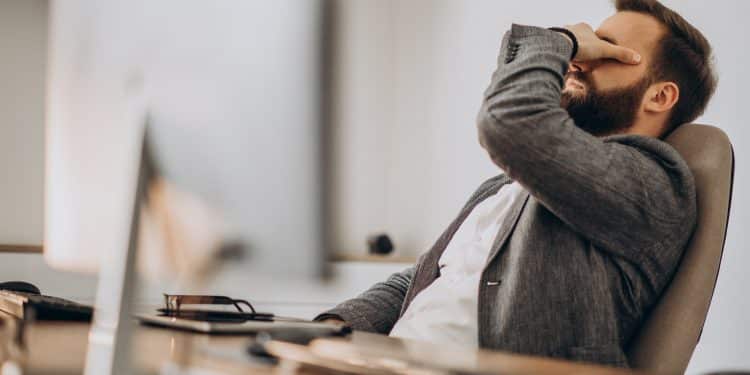 Homem de barba sentado em uma cadeira de escritório com a mão no rosto em expressão de bloqueio mental, representando o esquecimento momentâneo de palavras