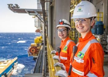 Duas profissionais sorrindo com uniforme laranja, capacetes brancos e óculos de segurança da Petrobras em plataforma offshore com o mar ao fundo