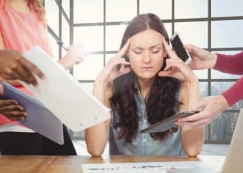 Mulher com olhos fechados pressionando as têmporas, cercada por mãos com celulares, papéis e tablets em ambiente de escritório