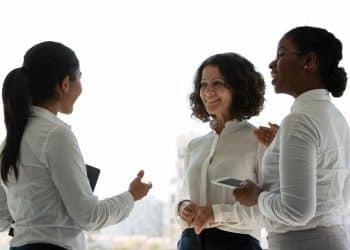 Três mulheres em traje social conversando e sorrindo em ambiente corporativo iluminado pela luz natural