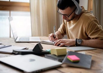 Estudante de fone de ouvido escreve em caderno ao lado de notebook em uma mesa de estudos, representação da rotina de preparação para o Enem com apoio de ferramentas de inteligência artificial.
