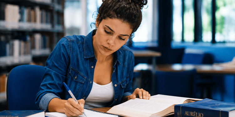 Jovem de cabelos cacheados presos faz anotações em caderno enquanto consulta livro aberto, com Dicionário Aurélio sobre mesa em biblioteca.