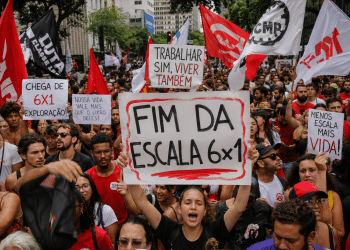 Manifestantes erguem cartazes pedindo fim da escala 6x1 na Avenida Paulista, com bandeiras vermelhas e brancas ao fundo.