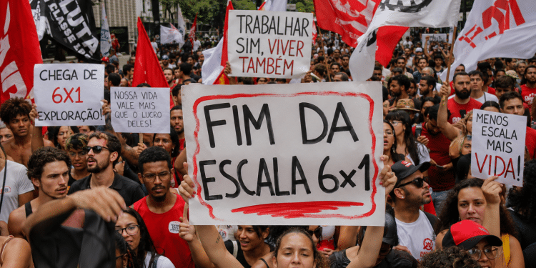 Manifestantes erguem cartazes pedindo fim da escala 6x1 na Avenida Paulista, com bandeiras vermelhas e brancas ao fundo.