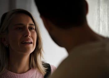 Mulher loira de cabelos lisos expressa angústia durante conversa tensa com homem visto de costas em ambiente com cortinas claras ao fundo