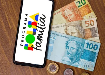 Celular exibindo logotipo do Programa Bolsa Família ao lado de cédulas de cem, cinquenta e vinte reais sobre mesa de madeira