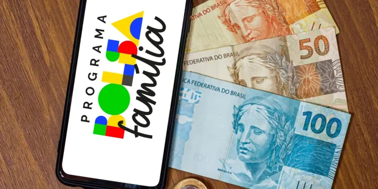Celular exibindo logotipo do Programa Bolsa Família ao lado de cédulas de cem, cinquenta e vinte reais sobre mesa de madeira