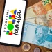Celular exibindo logotipo do Programa Bolsa Família ao lado de cédulas de cem, cinquenta e vinte reais sobre mesa de madeira