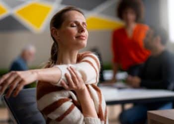 Mulher de olhos fechados alonga o braço e respira com tranquilidade em ambiente de trabalho.