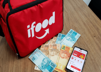 Bolsa térmica vermelha do iFood ao lado de notas de real e um celular com o aplicativo aberto sobre uma mesa