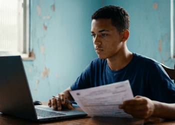 Jovem usa um notebook e segura um documento à mesa e, ao redor, um cômodo residencial de paredes azuis