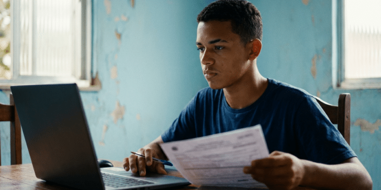 Jovem usa um notebook e segura um documento à mesa e, ao redor, um cômodo residencial de paredes azuis
