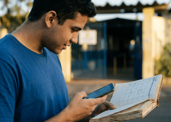 Jovem consulta calendário impresso e celular em frente a portão de escola pública