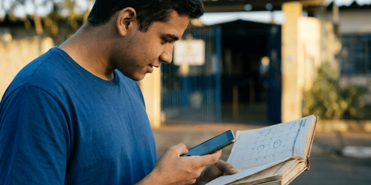 Jovem consulta calendário impresso e celular em frente a portão de escola pública
