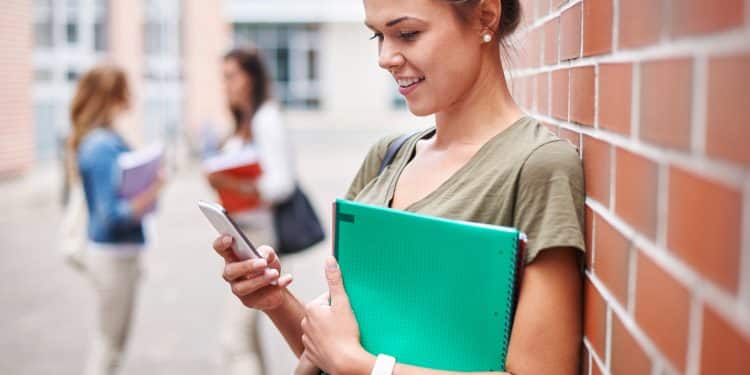 Estudante encostada em parede de tijolos consulta o celular enquanto segura caderno verde em pátio de instituição de ensino