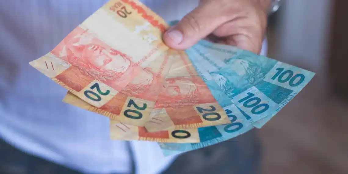 Homem segurando cédulas de real em referência ao pagamento de R$ 2.431,50 do INSS.
