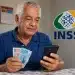 Aposentado brasileiro mexendo no celular e segurando notas de real, representando a consulta do 13º salário antecipado do INSS.