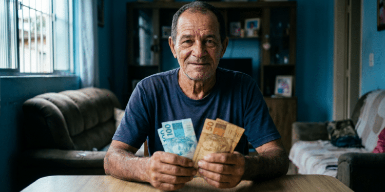Homem idoso segura notas de real sentado à mesa e, ao redor, a sala de uma casa com sofás e estante