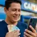 Homem consulta o celular em frente à agência da CAIXA enquanto segura notas de real, representando a liberação antecipada do Saque-Aniversário.