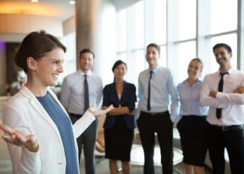 Executiva de blazer branco gesticula enquanto fala para cinco colaboradores sorridentes em lobby empresarial iluminado, simbolizando o curso Líder e Gestor de Equipes do Pensar Cursos.
