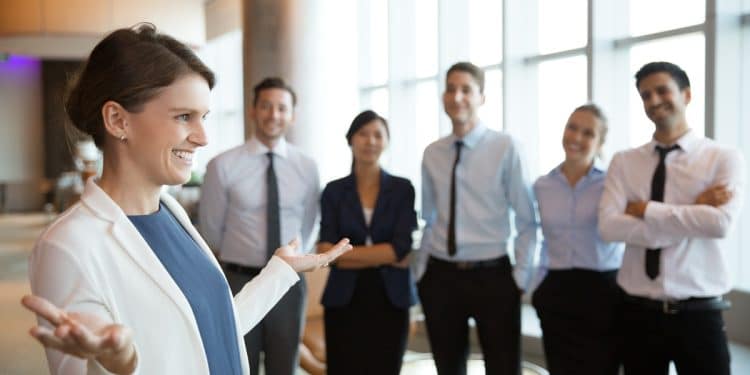 Executiva de blazer branco gesticula enquanto fala para cinco colaboradores sorridentes em lobby empresarial iluminado, simbolizando o curso Líder e Gestor de Equipes do Pensar Cursos.