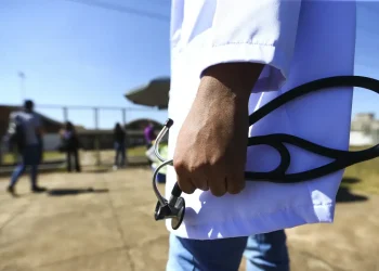 Médico de jaleco branco segurando um estetoscópio ao ar livre, em referência às vantagens de seguir a carreira de Medicina.
