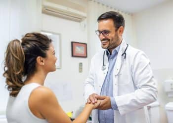 Médico de jaleco branco e estetoscópio sorri e segura as mãos de paciente em consultório médico iluminado, simbolizando o título de doutor dado à profissão.