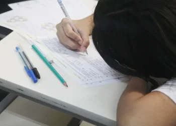 Jovem estudante concentrada escreve em folha de prova sobre carteira escolar em imagem ilustrativa sobre as novas regras do Enem 2026